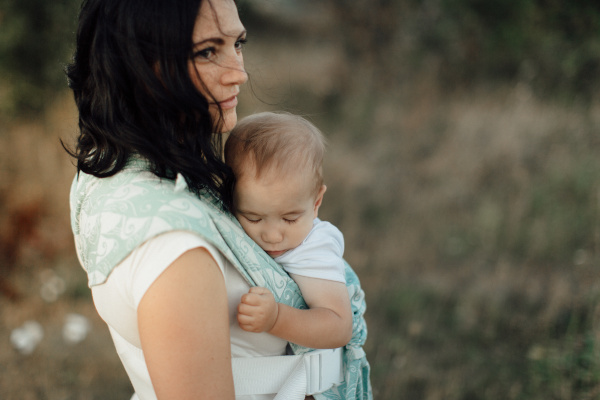 Mother wearing her sleeping child in carrier. Portrait of woman carrying her little baby in sling in the fields on summer day.